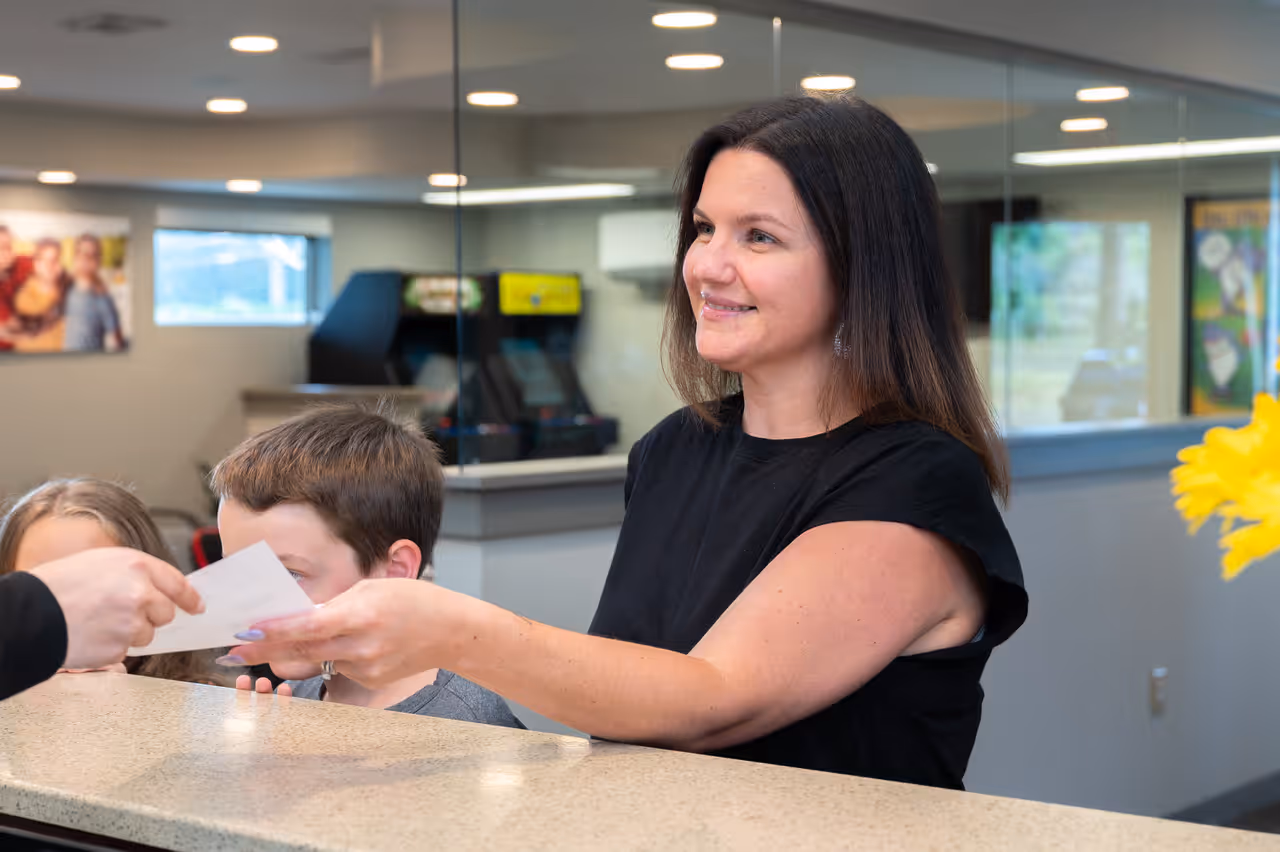 Smiling woman at reception desk handing a card to a person across the counter in an office setting.