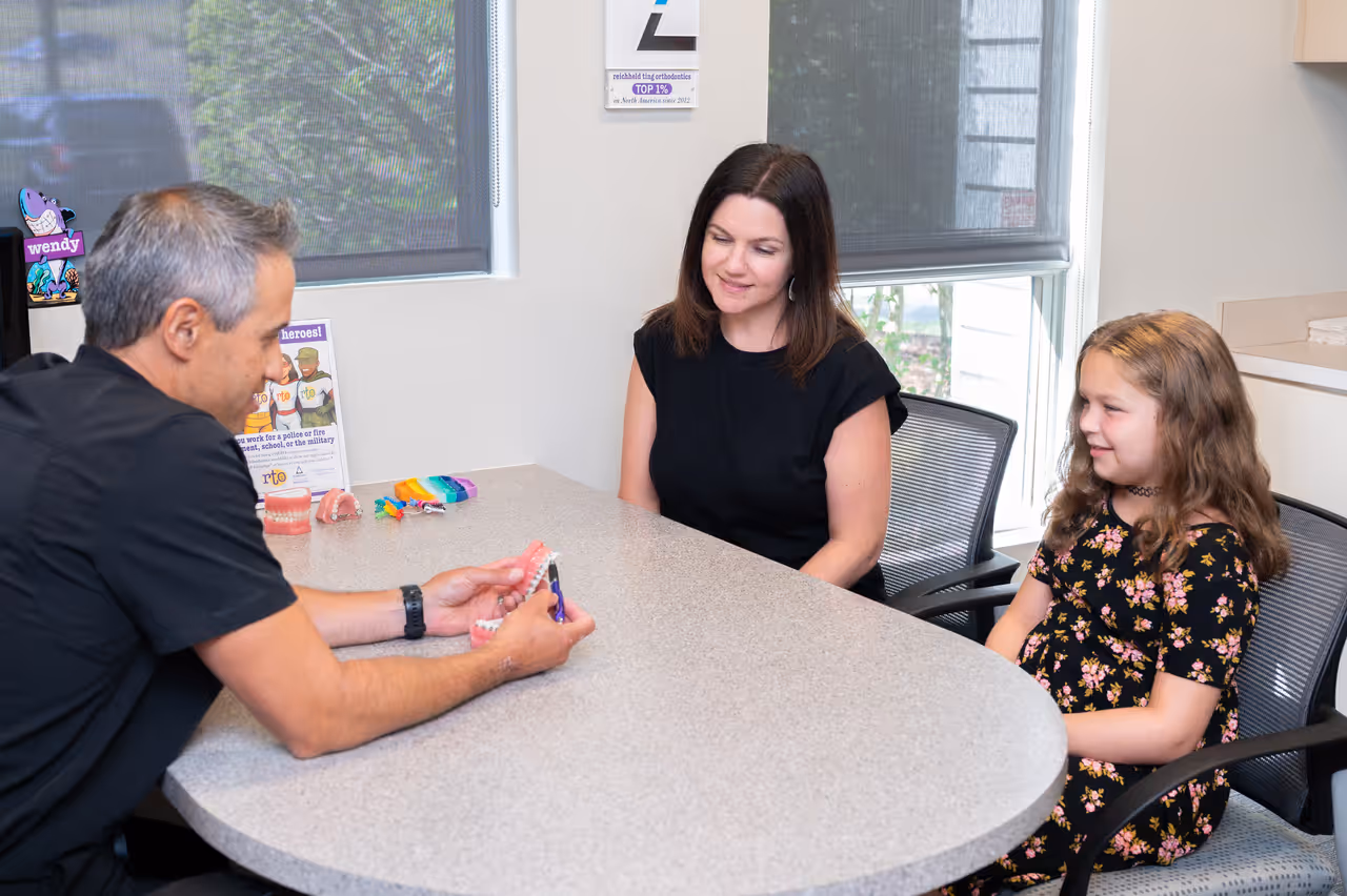 Orthodontist explaining braces to a woman and a young girl sitting at a consultation table.