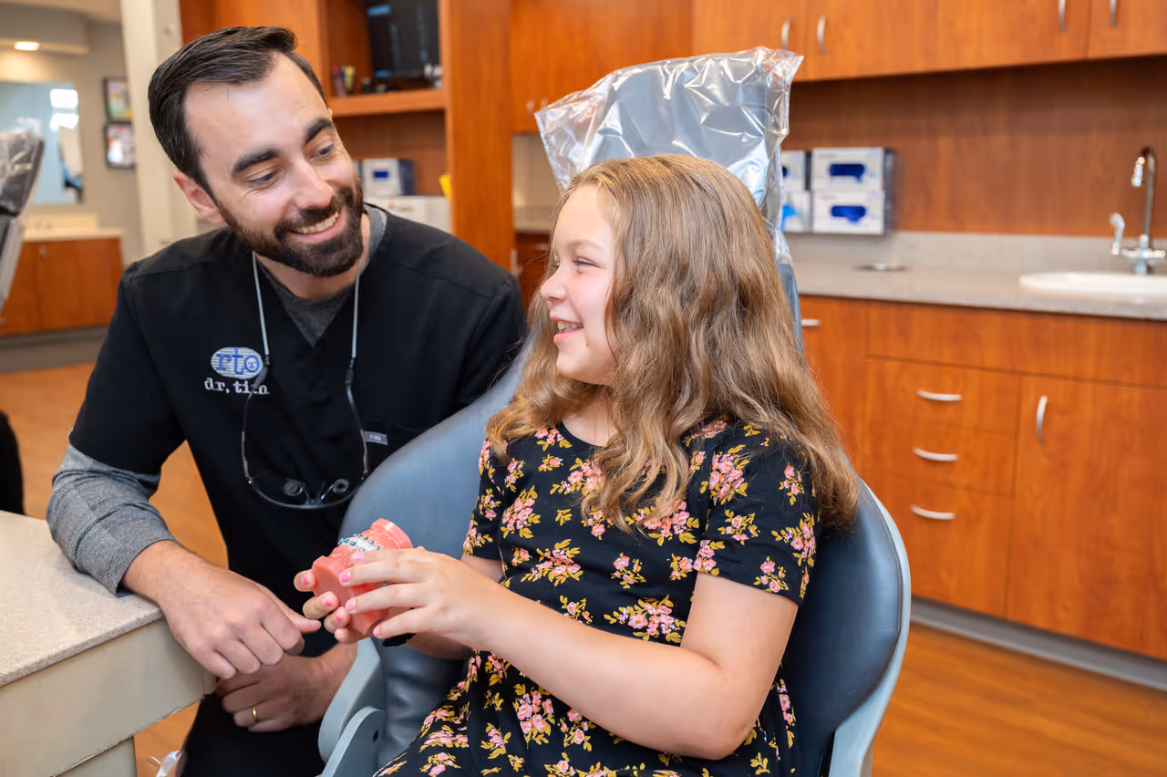 Orthodontist smiling and talking to a girl holding a dental model in a clinic chair.
