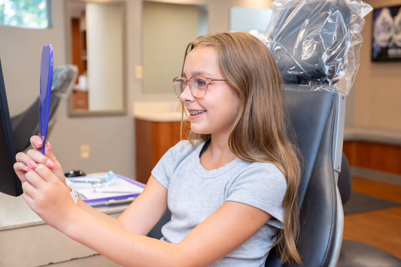 Smiling girl with braces and glasses sitting in an orthodontic chair looking into a handheld mirror.