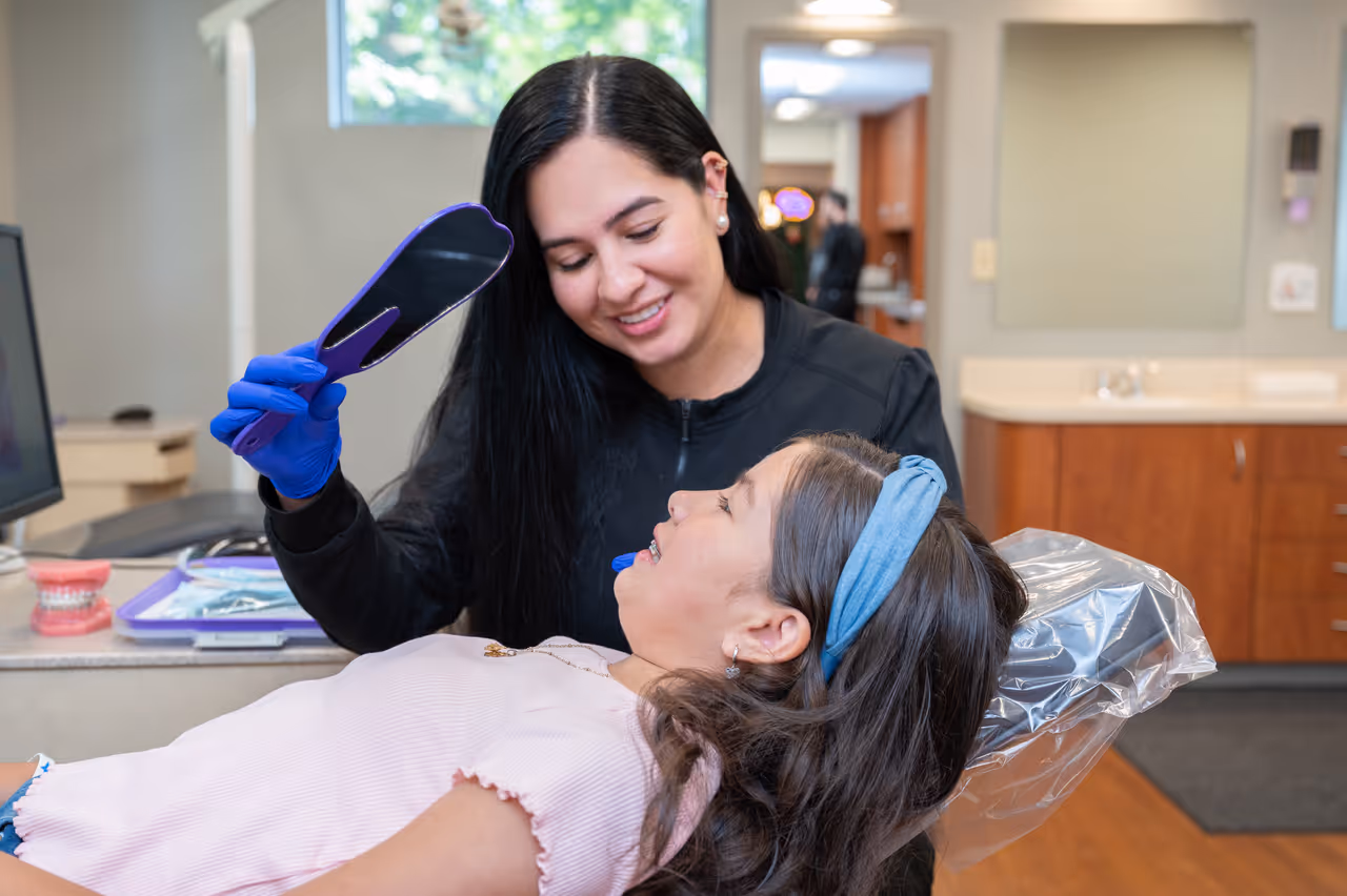 Orthodontist showing a smiling girl with braces her reflection in a handheld mirror at dental office.