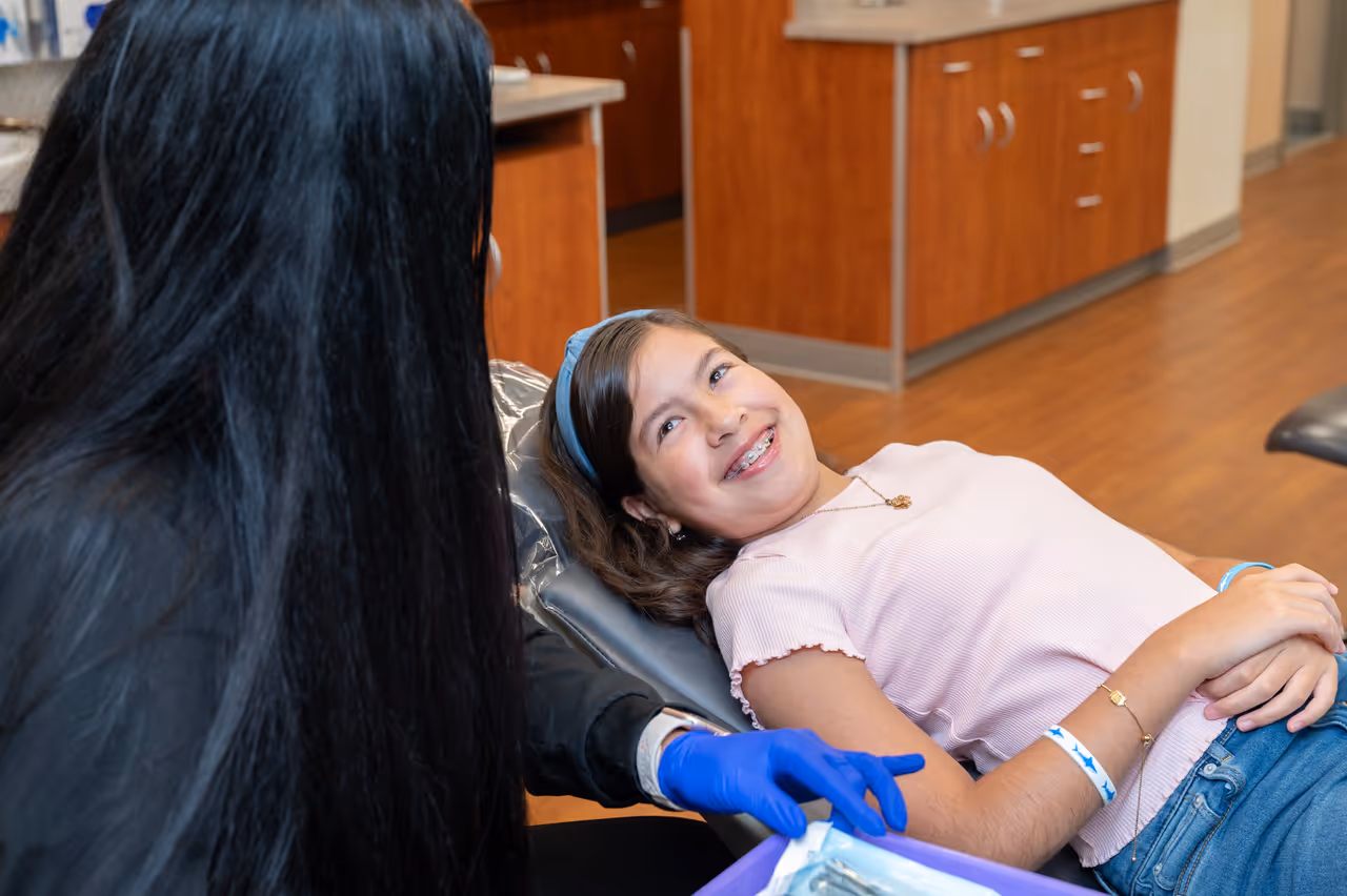 Smiling girl with braces lying in dentist chair talking to a dental professional wearing blue gloves.