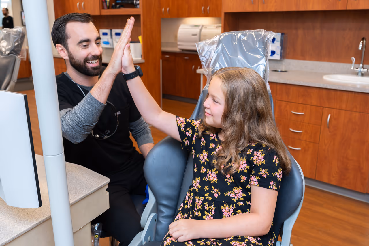 Smiling male dentist and young girl in floral dress sharing a high five in dental office.