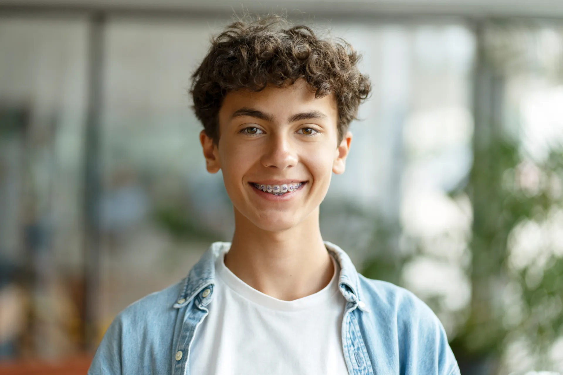 Smiling teenage boy with curly hair wearing braces, a white shirt, and a denim jacket.