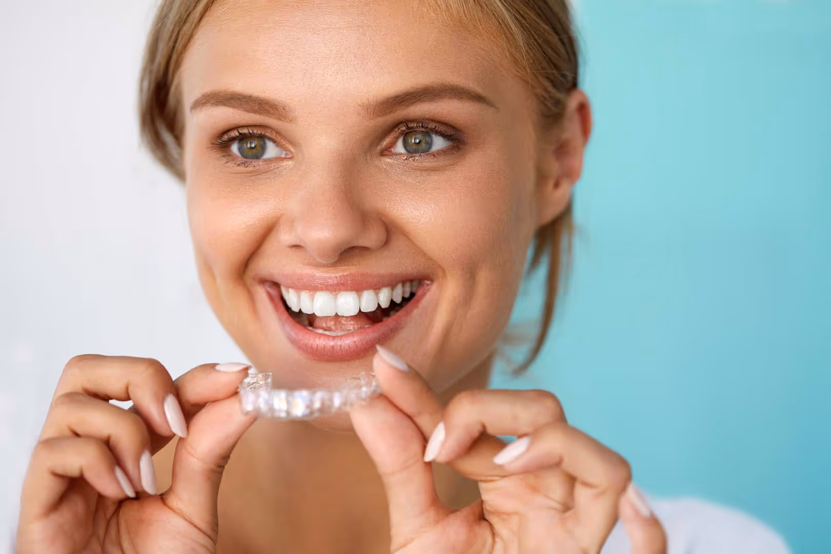Smiling woman holding a clear dental aligner with well-manicured nails against blue background.