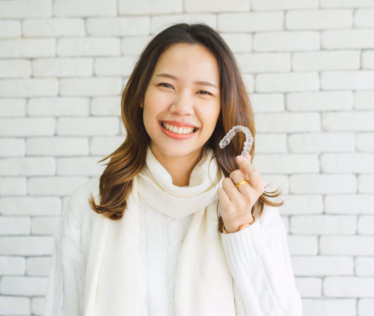 Smiling woman in white sweater holding a clear orthodontic aligner against a white brick wall.