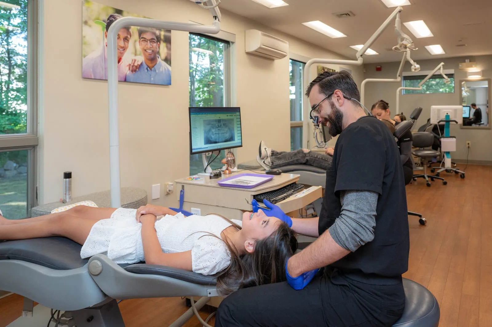Dentist wearing magnifying glasses and blue gloves examining a smiling girl lying on dental chair.