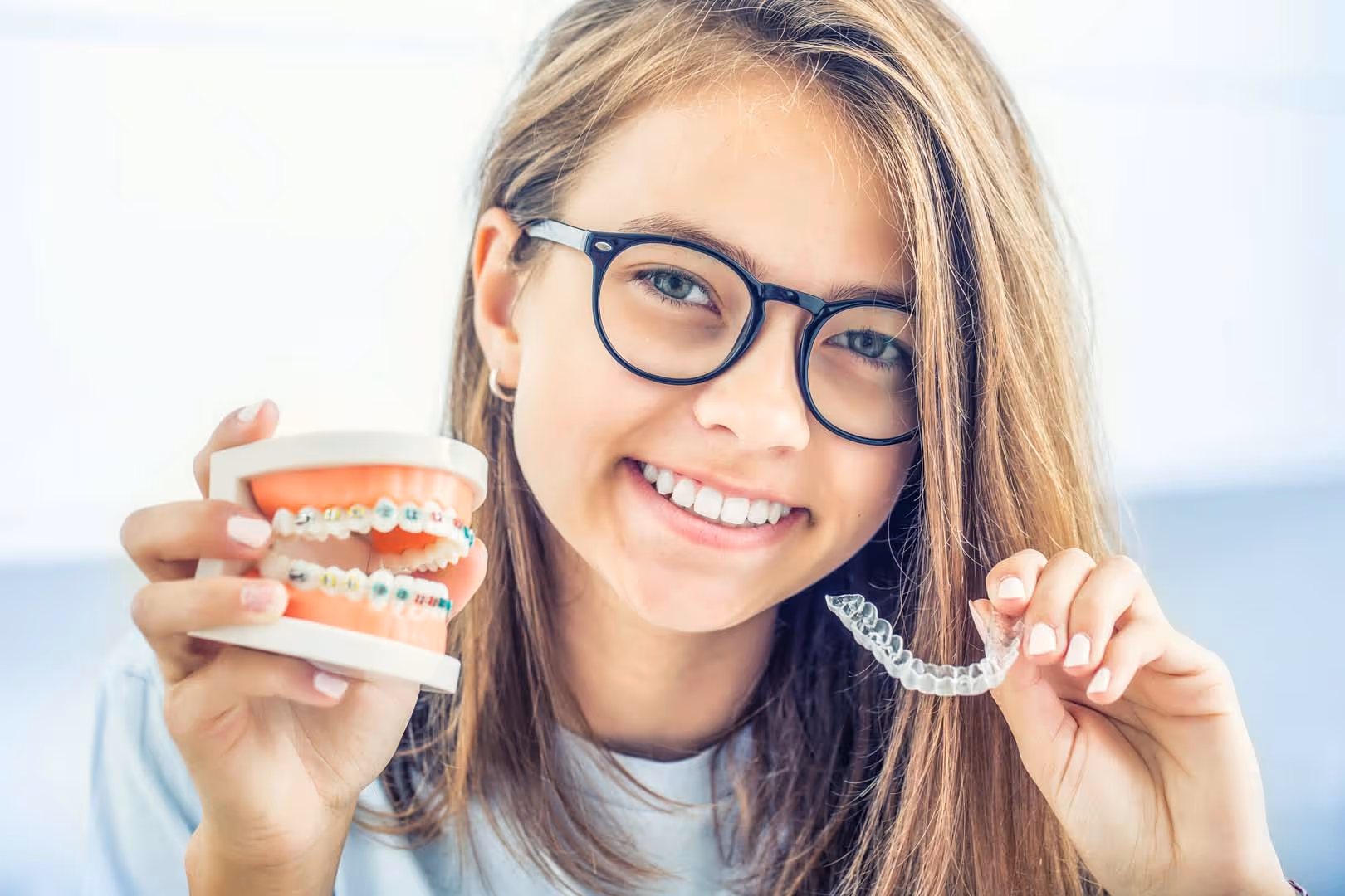 Smiling woman with glasses holding a braces model in one hand and a clear aligner in the other.