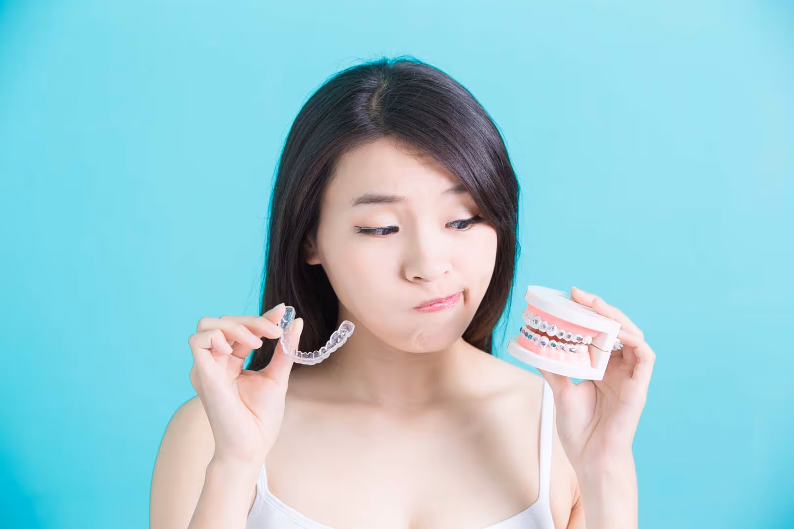 Young woman comparing clear aligner and braces model with uncertain expression on blue background.