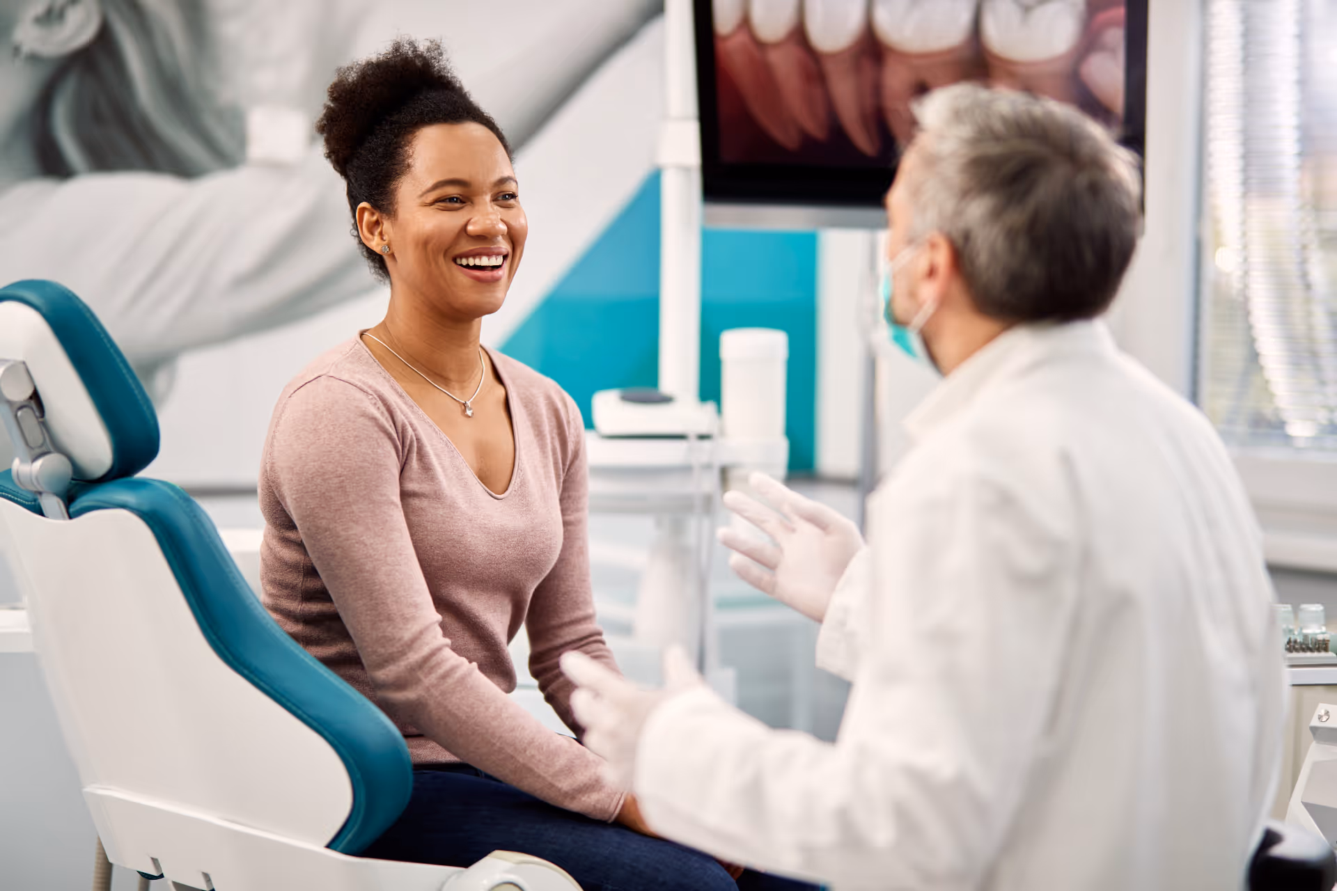 Smiling woman sitting in dental chair talking with a male dentist wearing gloves and mask.