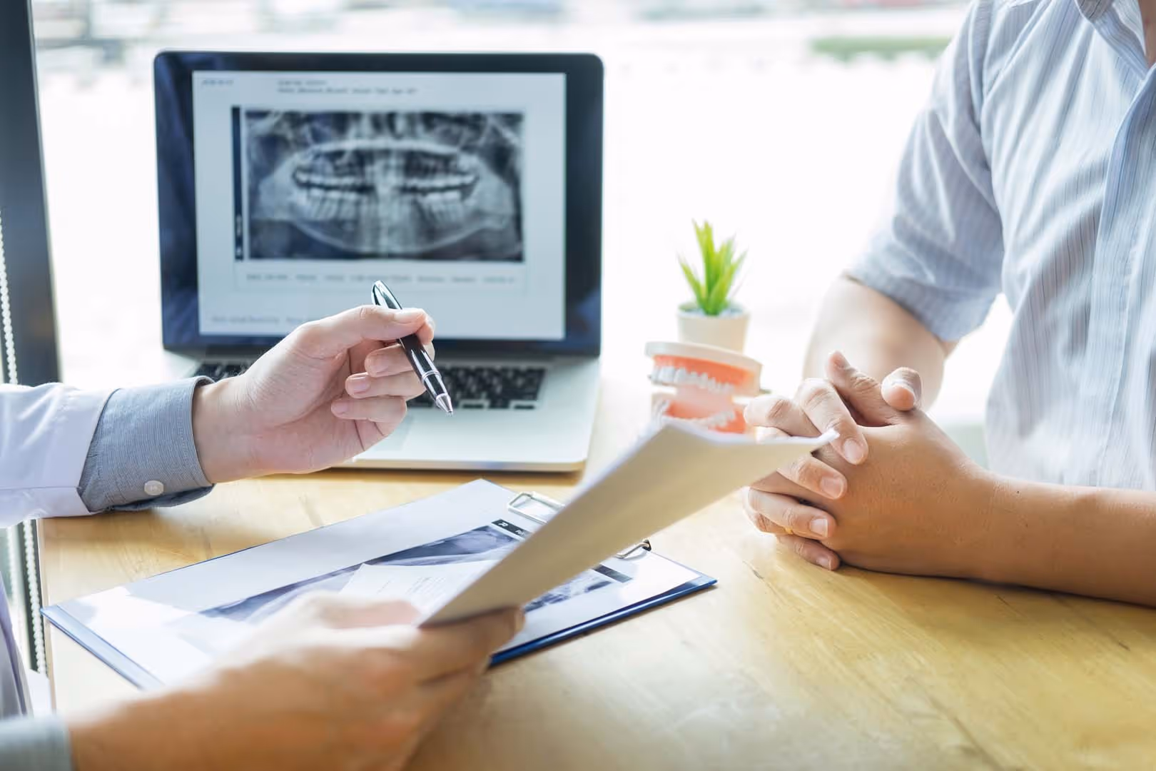 Dentist showing dental X-ray on laptop and holding paper while patient listens with folded hands.