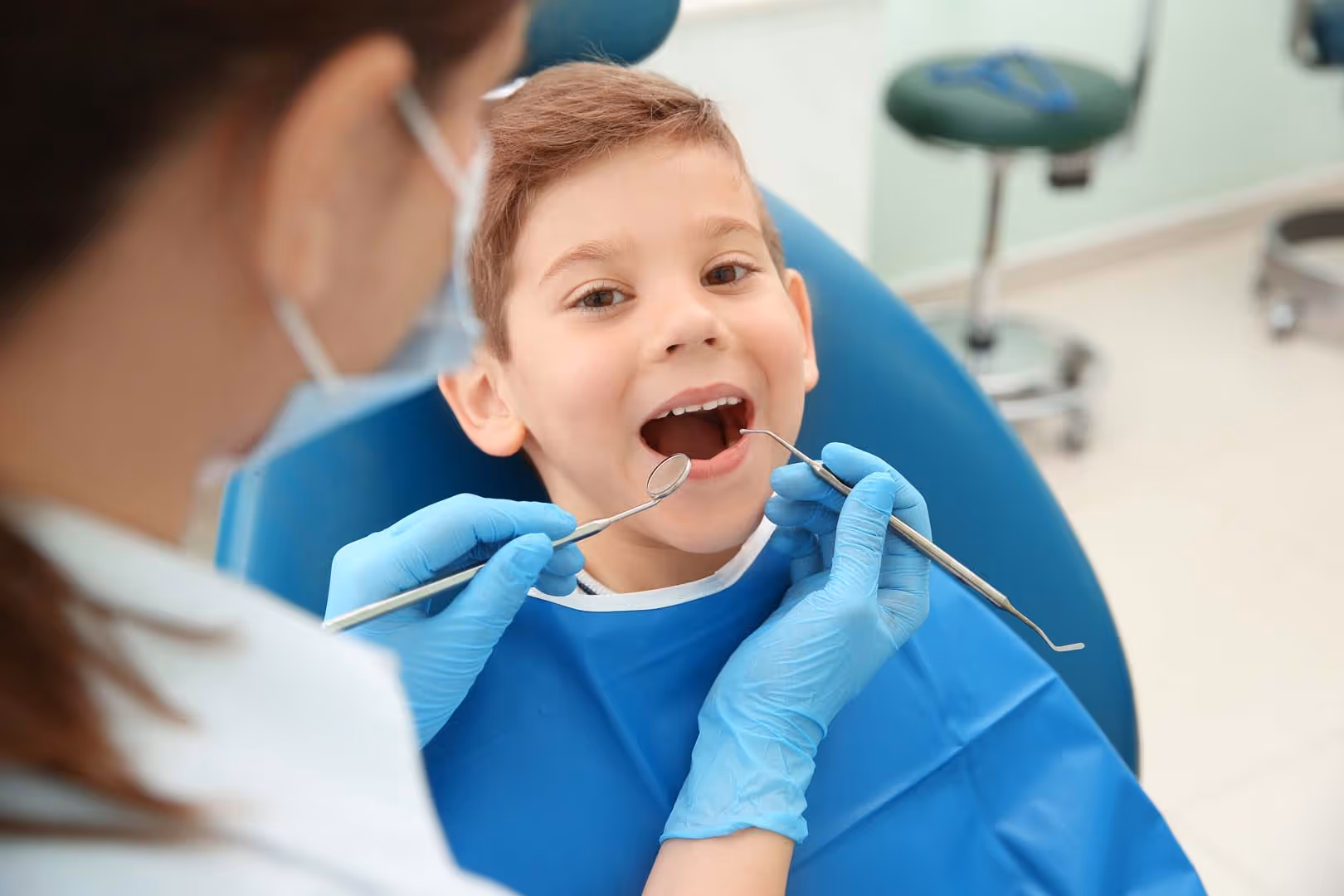 Dentist wearing gloves examines young boy's teeth using dental mirror and probe in clinic chair.