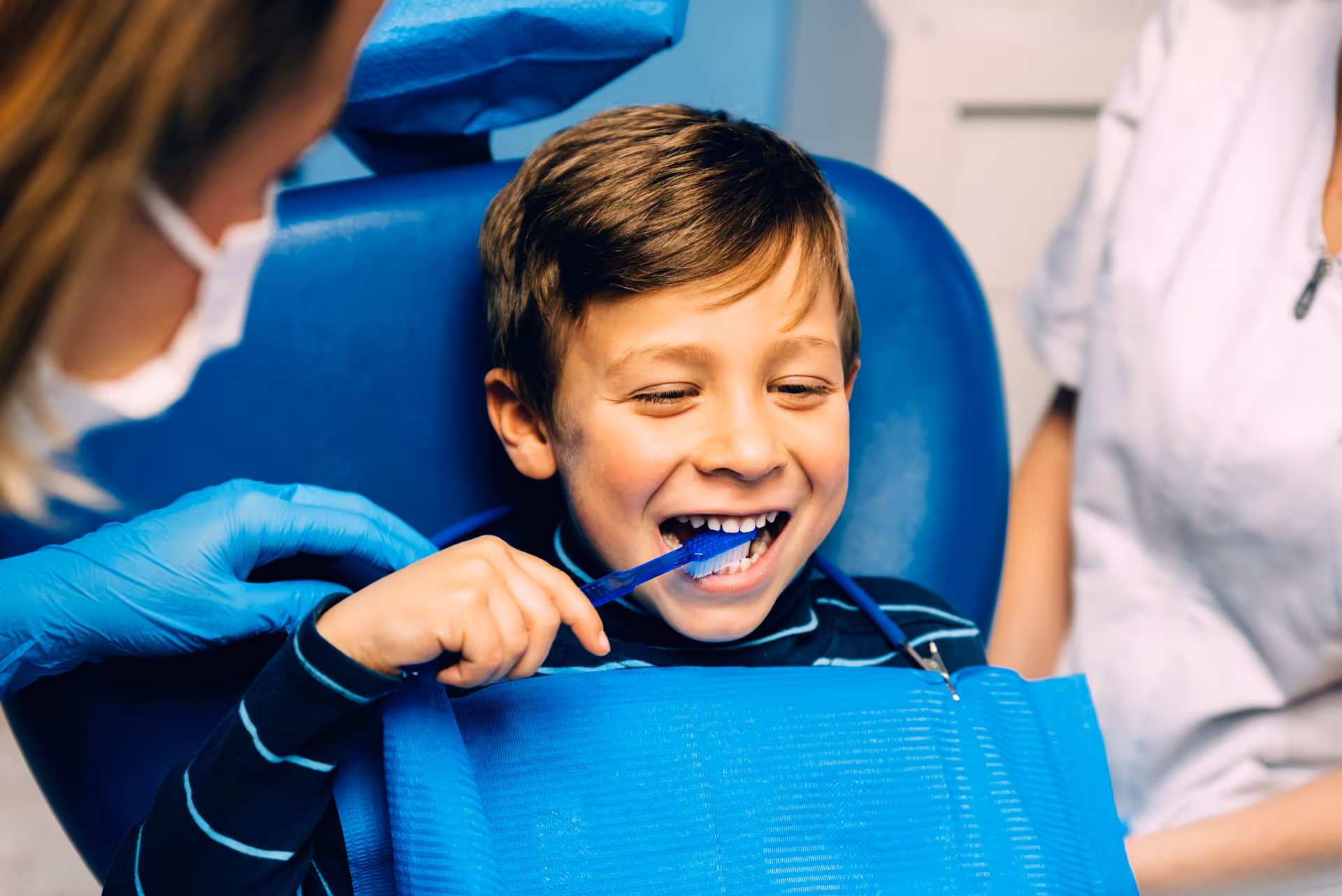 Boy in dental chair smiling while holding a toothbrush with dentist's gloved hand nearby.