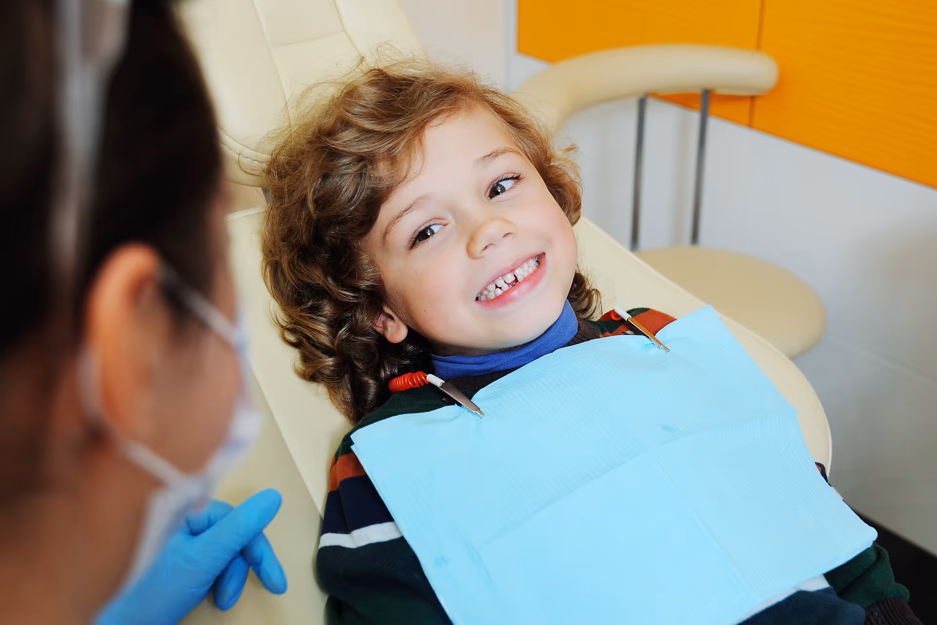 Smiling child wearing a dental bib sitting in a dentist chair talking to a masked dentist.