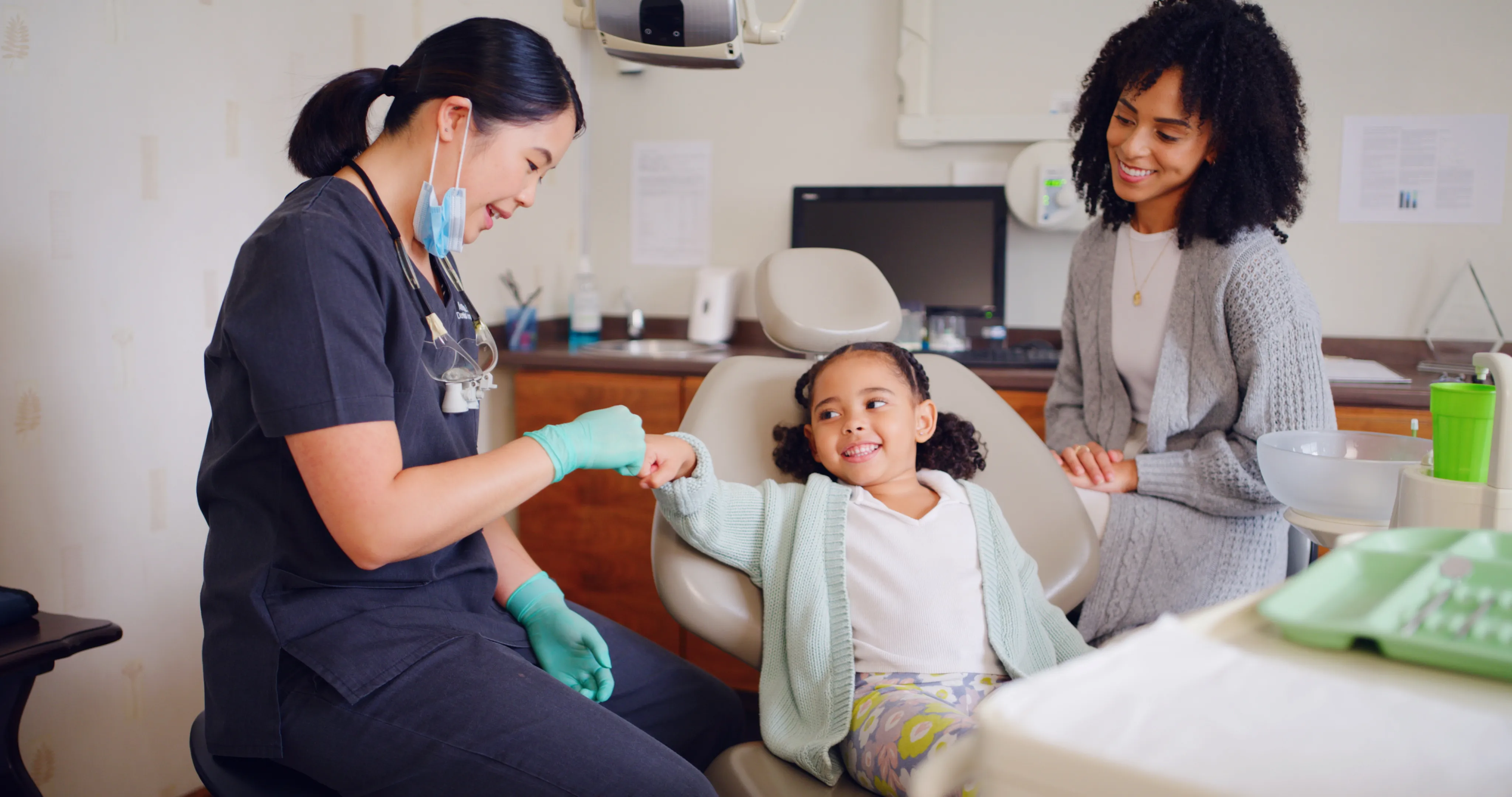 Dentist in gloves fist bumping smiling young girl seated in dental chair with her mother nearby.