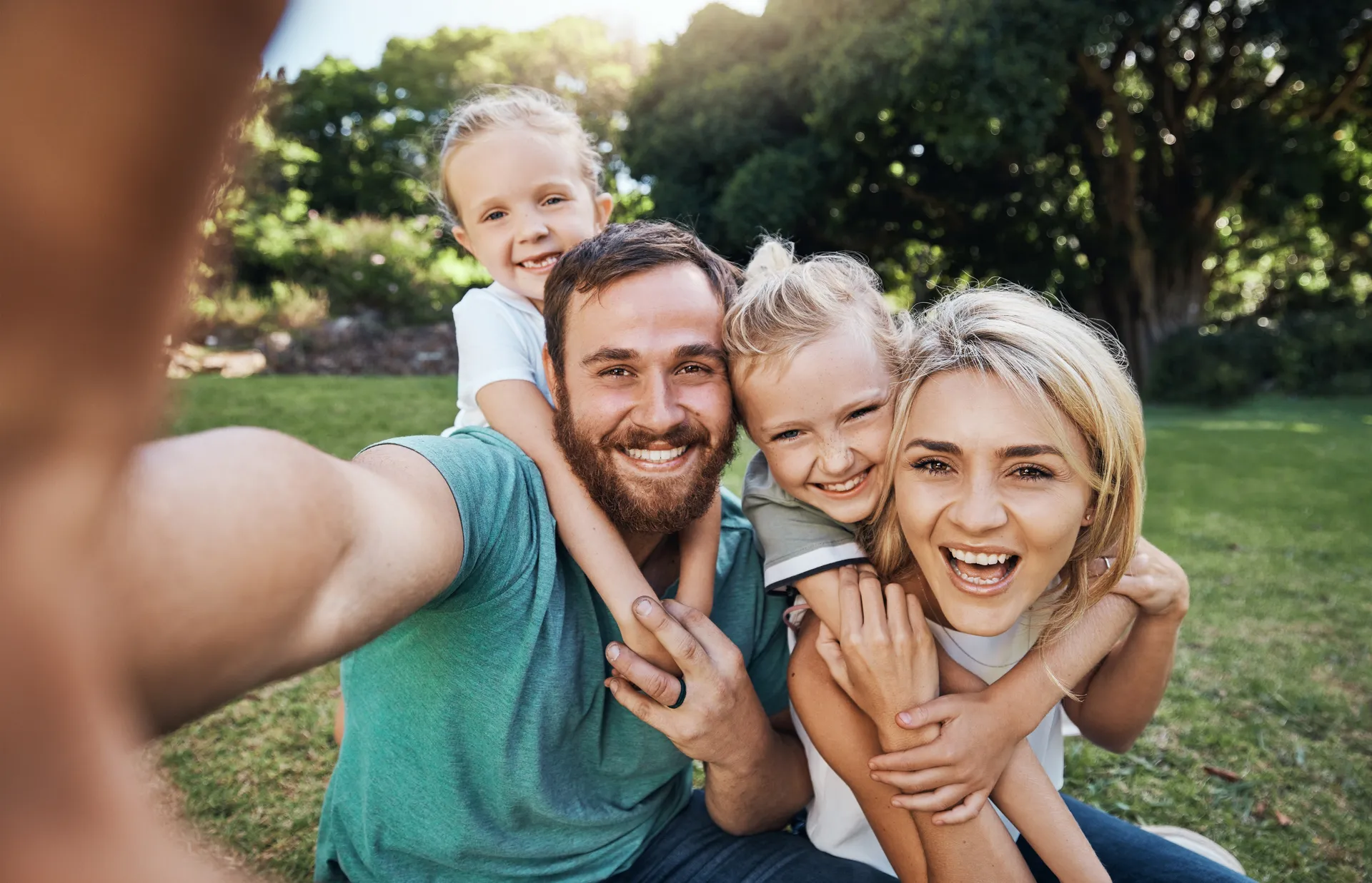 Smiling family of four sitting on grass outdoors taking a selfie in a sunny park.
