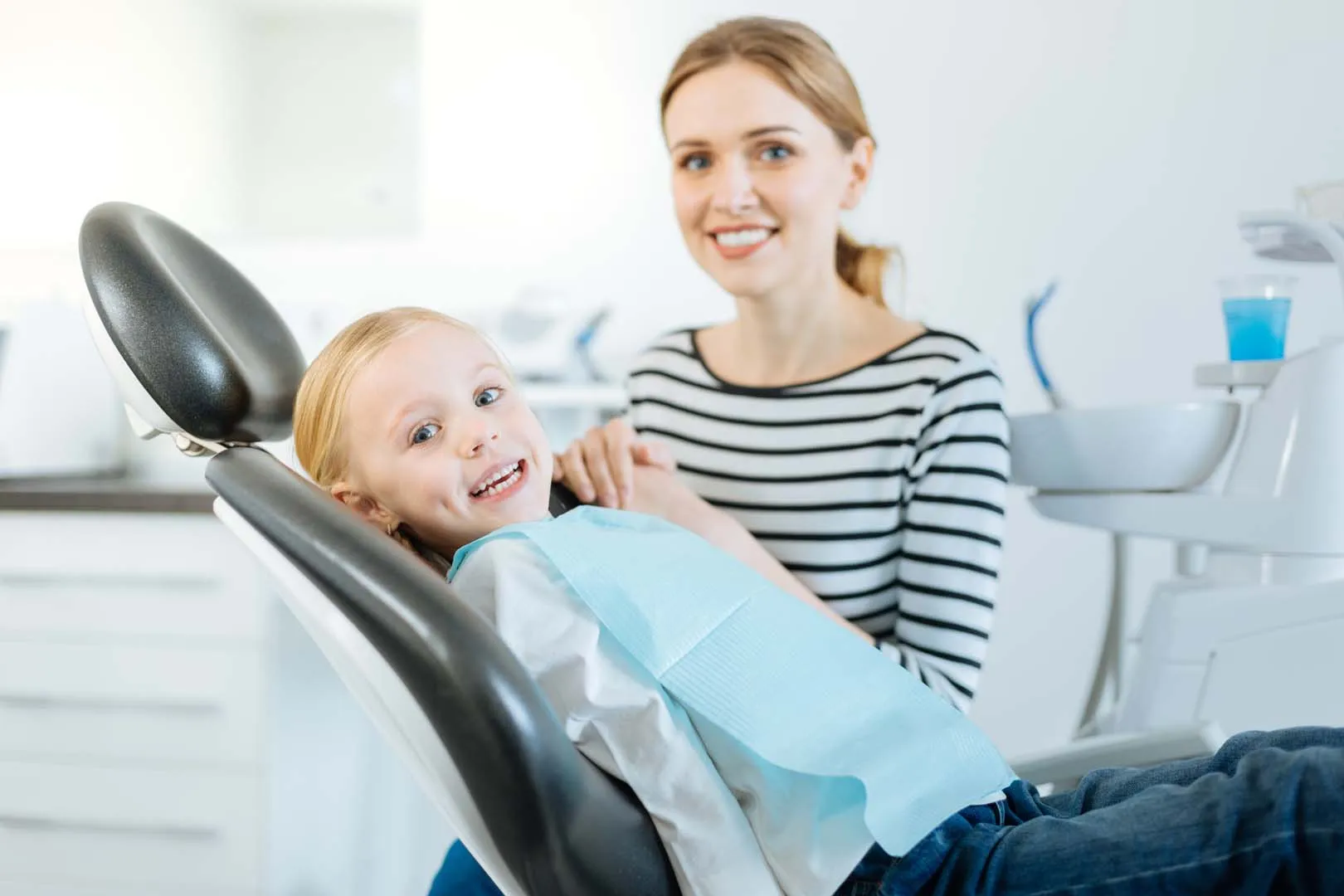 Smiling child sits in dental chair with blue bib while a woman in stripes supports from behind.
