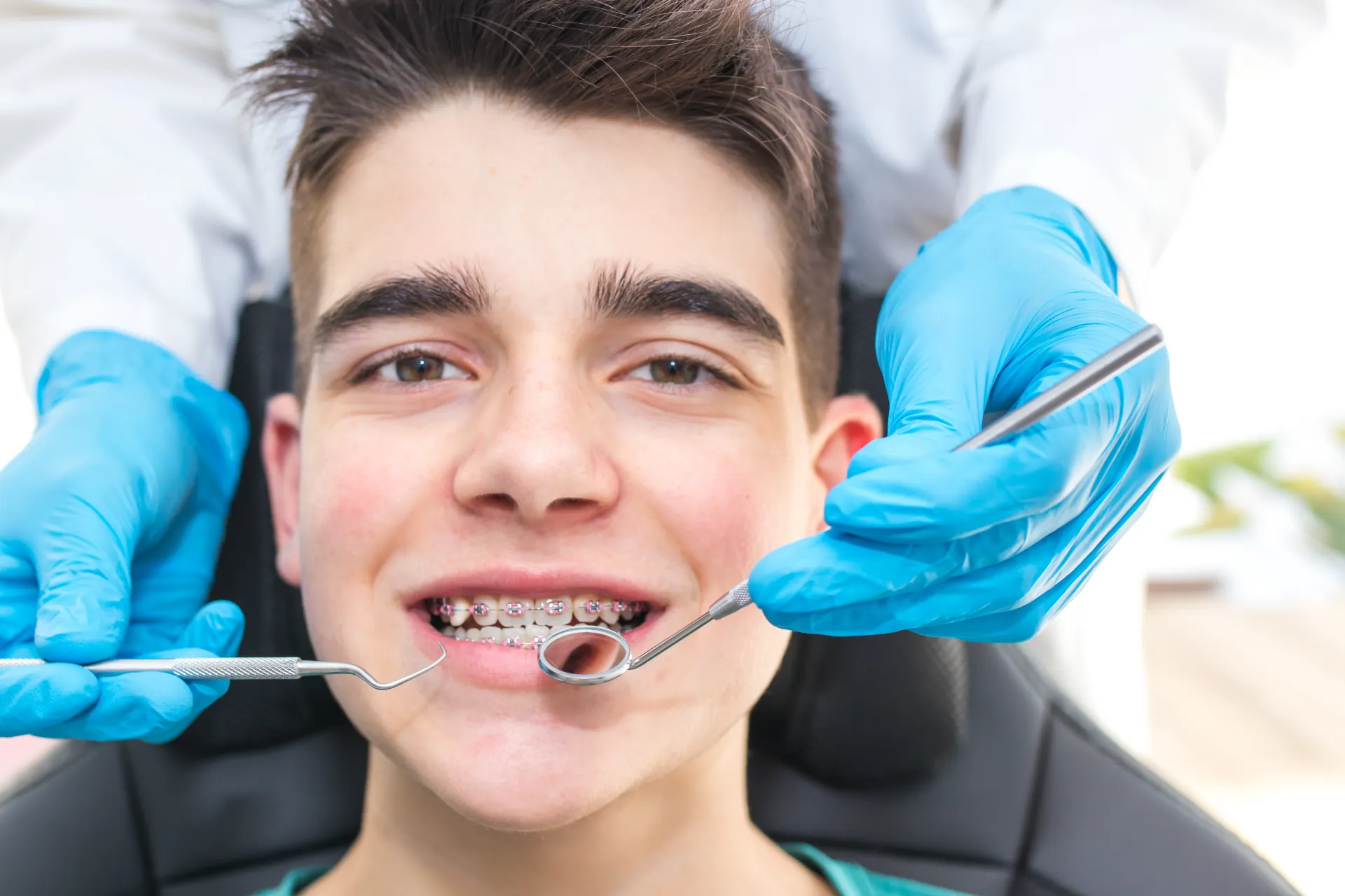 Close-up of a smiling teen with braces during a dental checkup with tools held by a gloved dentist.