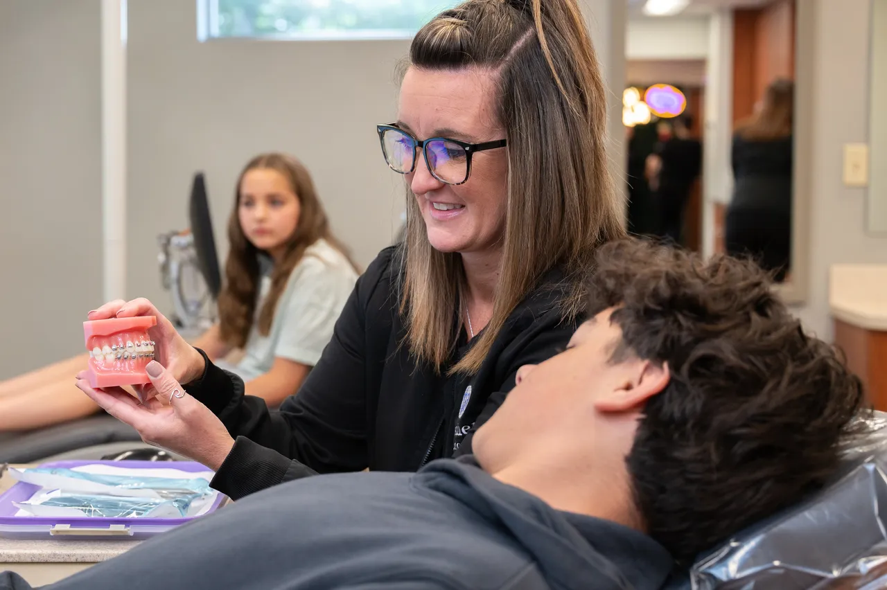 Orthodontist showing braces model to a teen patient in a dental clinic with another patient in background.