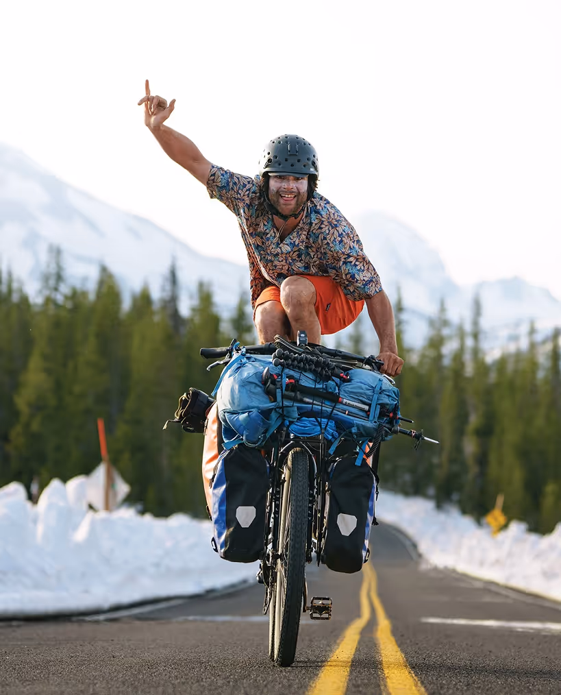 Stratton Matteson wearing helmet riding a loaded touring bike on a mountain road, making a hand gesture and smiling.