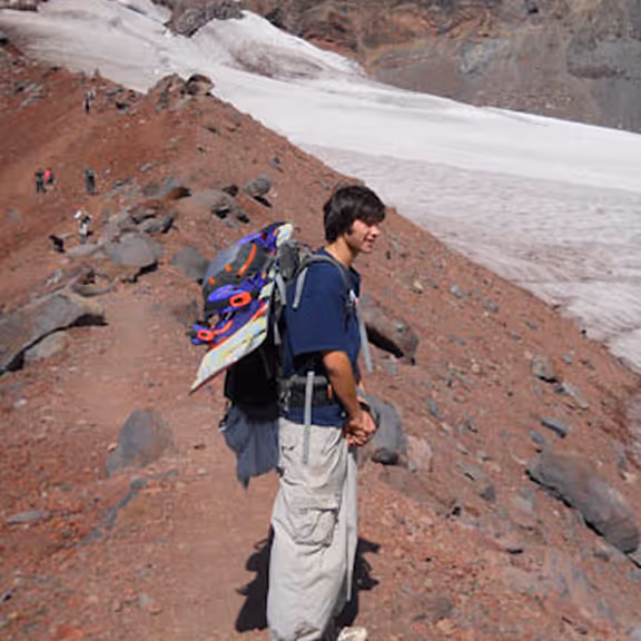 A teenage Stratton hiking a volcanic trail with a snowboard strapped to his backpack.