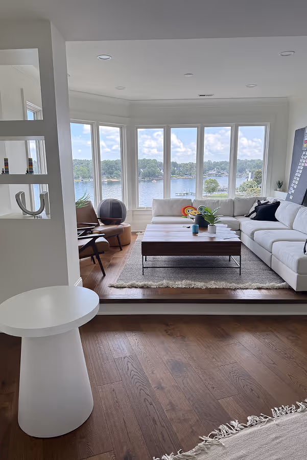 Living room with large windows overlooking a lake, white sectional sofa, wooden coffee table, and leather chair.