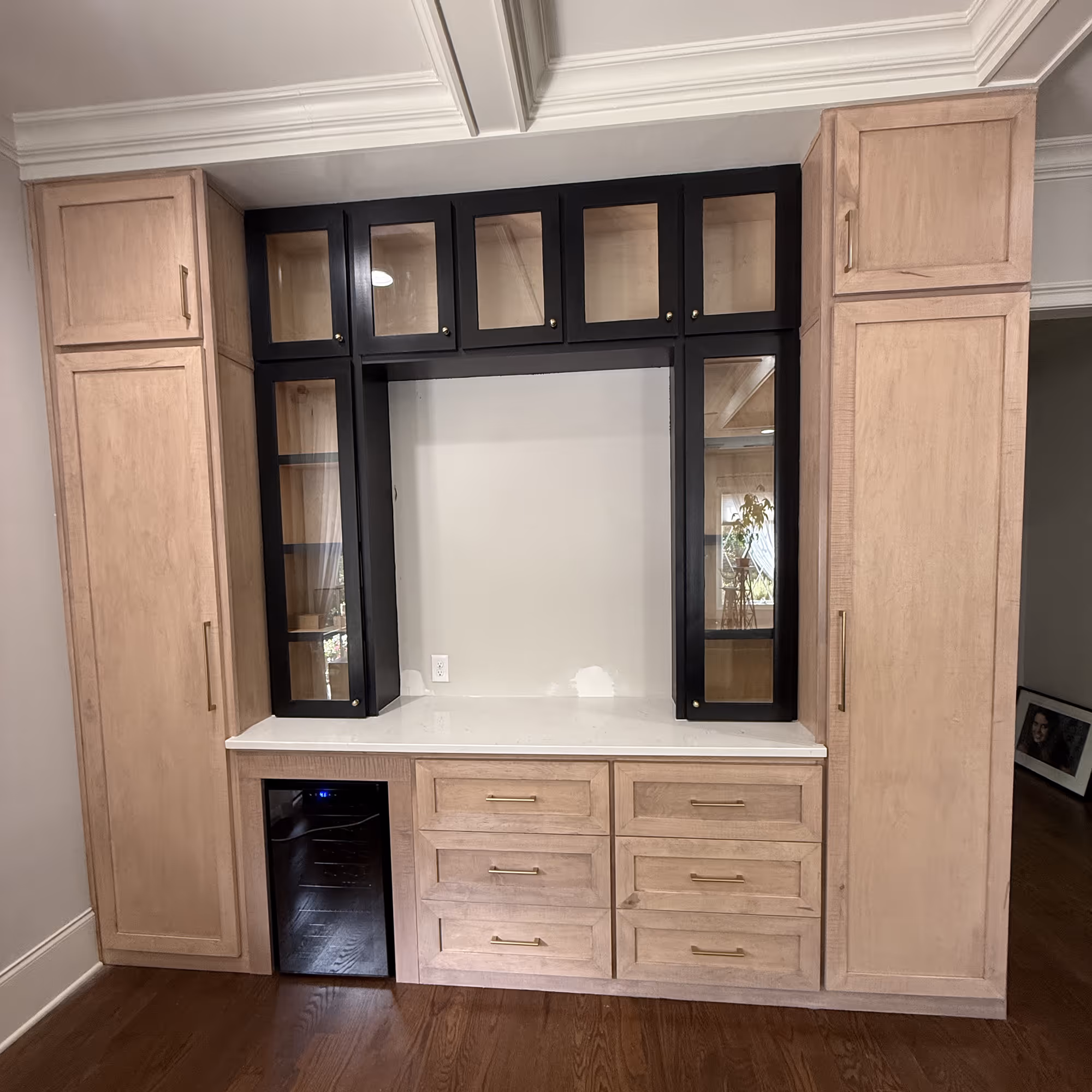 Custom light wood wall unit with black-framed glass cabinets, drawers, countertop, and wine fridge.