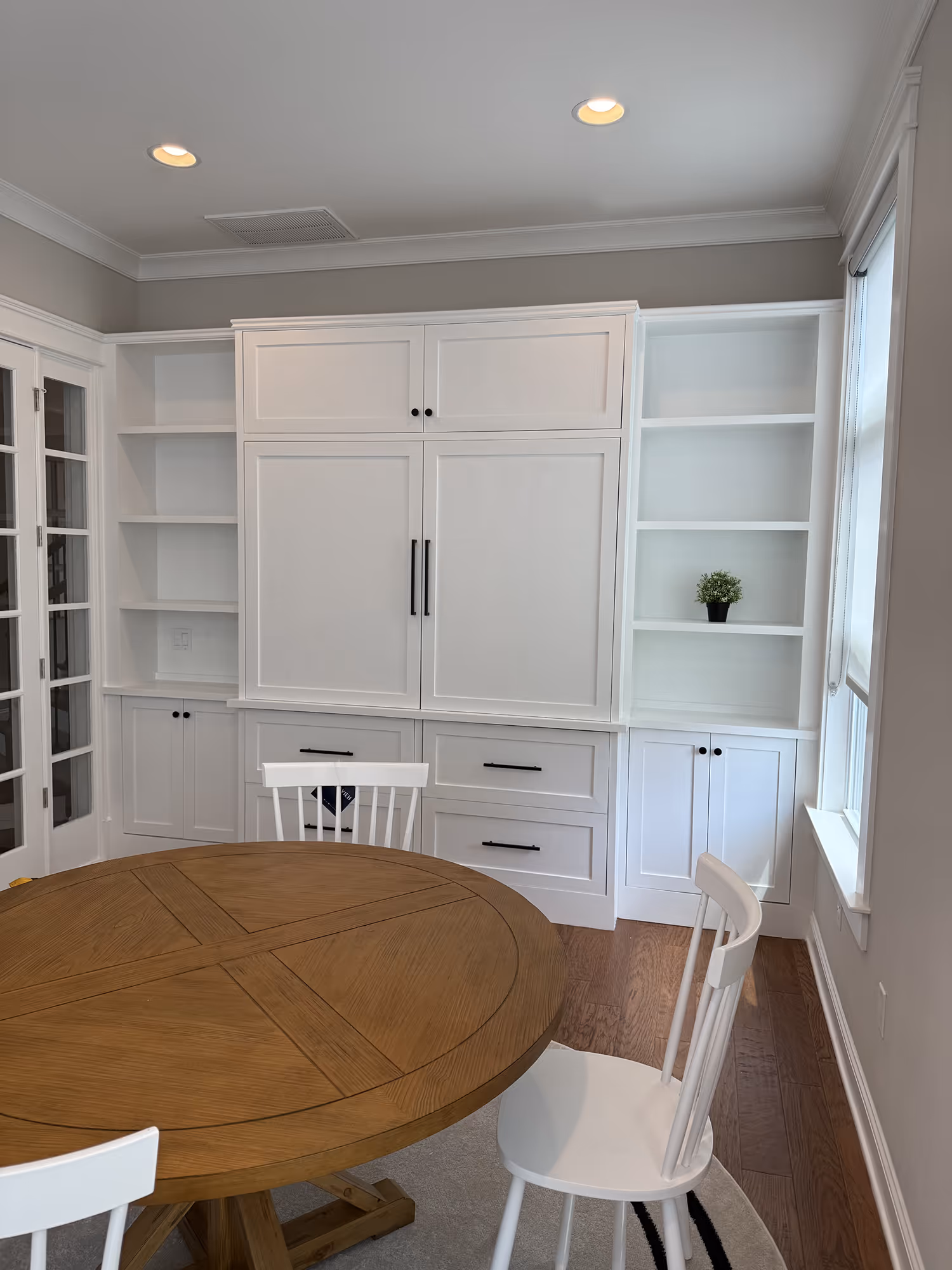 Dining area with round wooden table, white chairs, built-in white shelves, and a small potted plant.