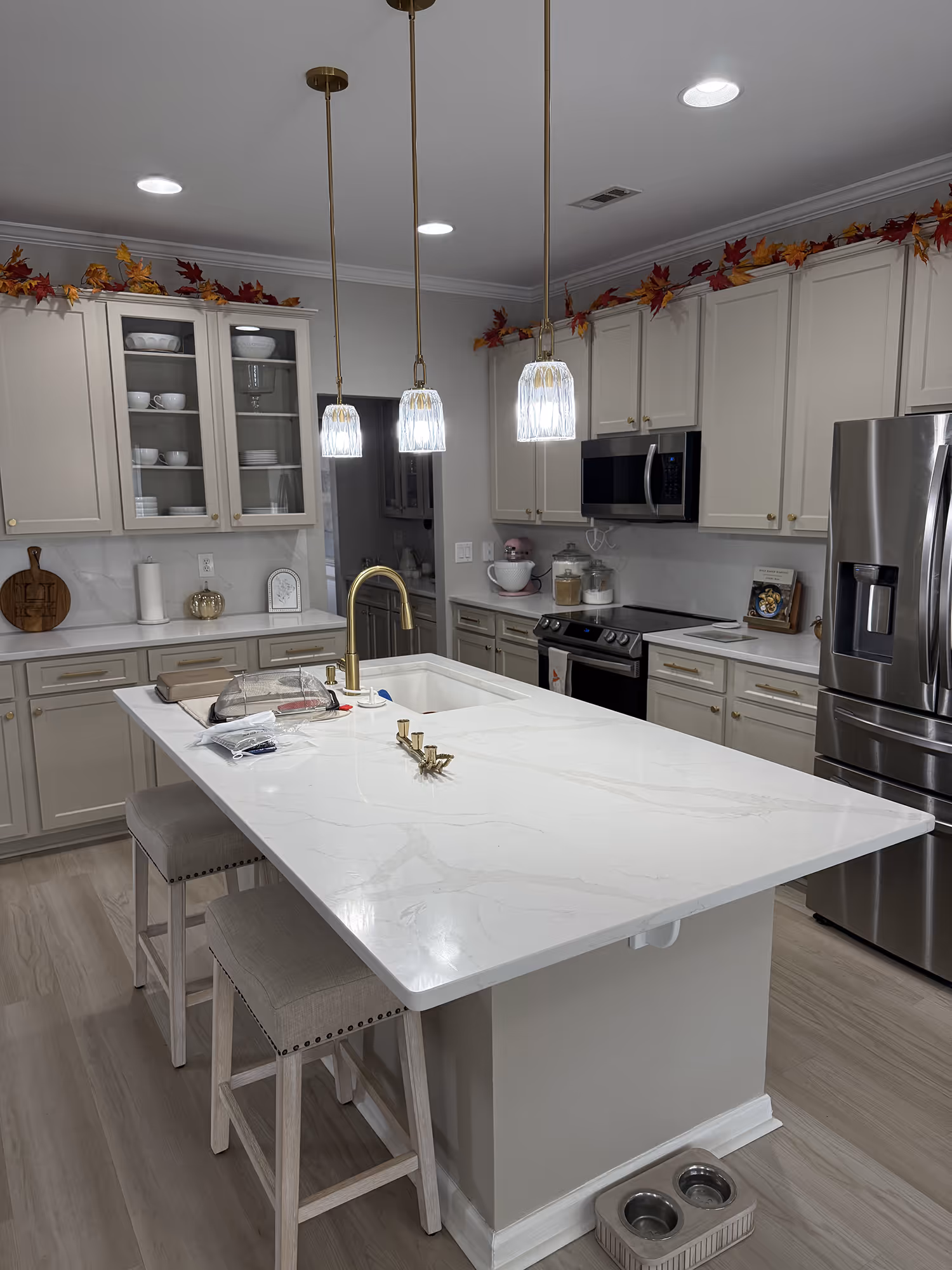 Modern kitchen with white marble island, beige cabinets, stainless appliances, and autumn leaf decor.