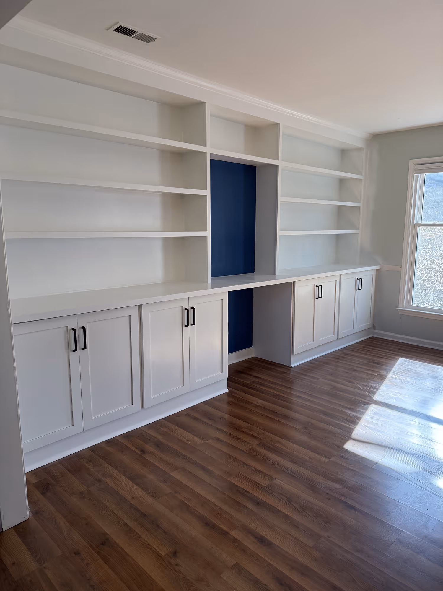 Empty white built-in shelves and cabinets with a blue back panel and wood floor in a sunlit room.