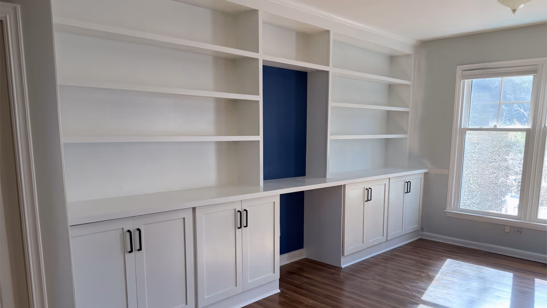 Empty white built-in shelves and cabinets with a blue back panel and wood floor in a sunlit room.