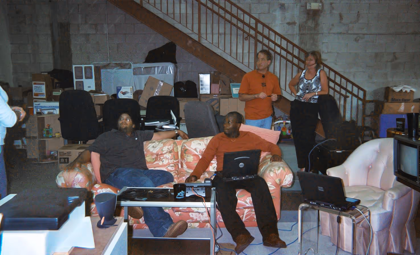 Four people in a cluttered basement office setup with laptops and mismatched furniture.