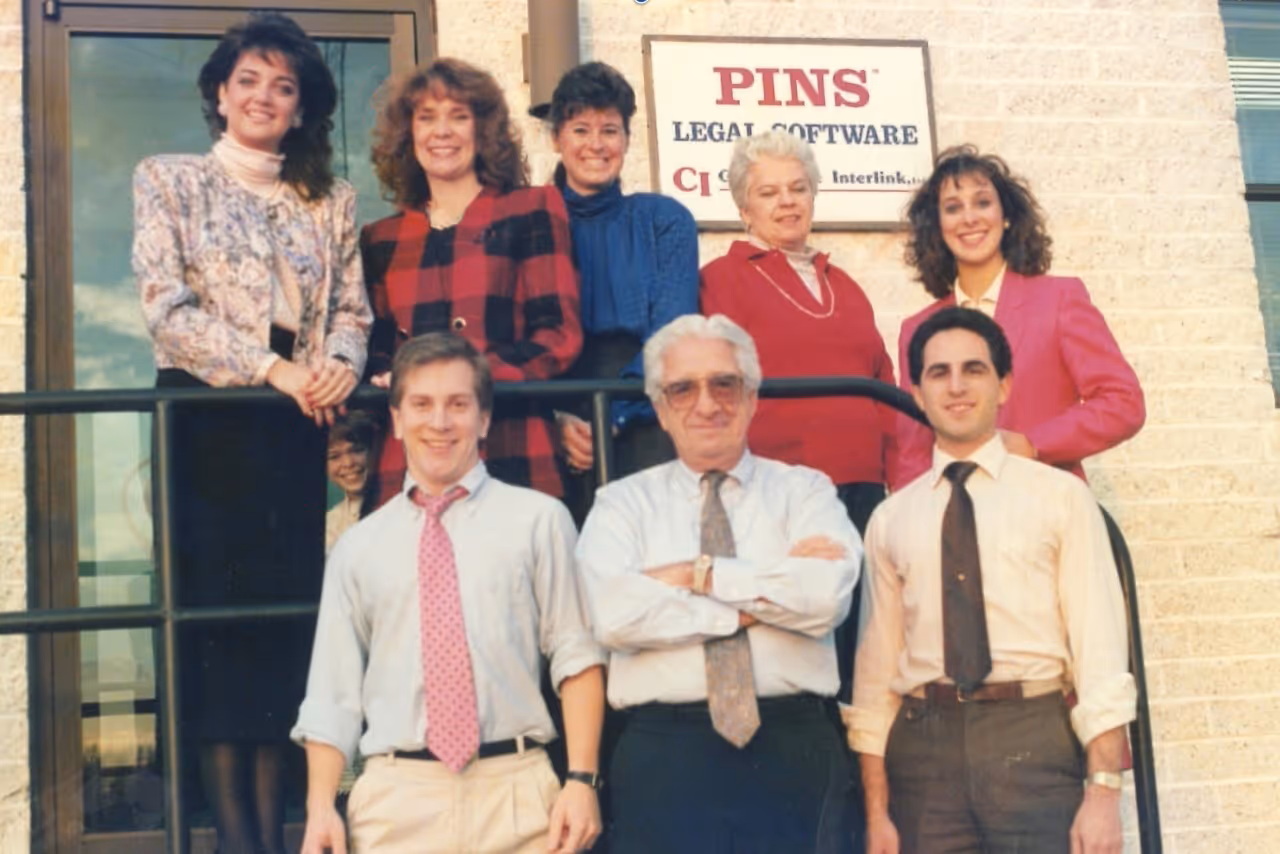 1980s group photo of eight PINS Legal Software staff members standing outside a building.