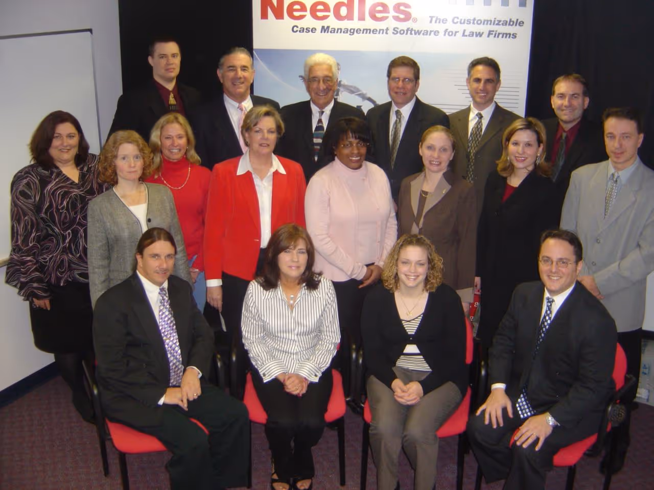 A group of 17 professionally dressed Assembly team members posing indoors with a banner behind them.