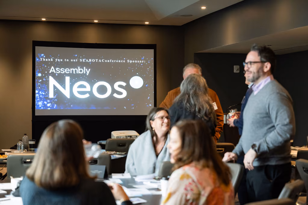 People socialize in a conference room with the Assembly Neos logo on a screen.