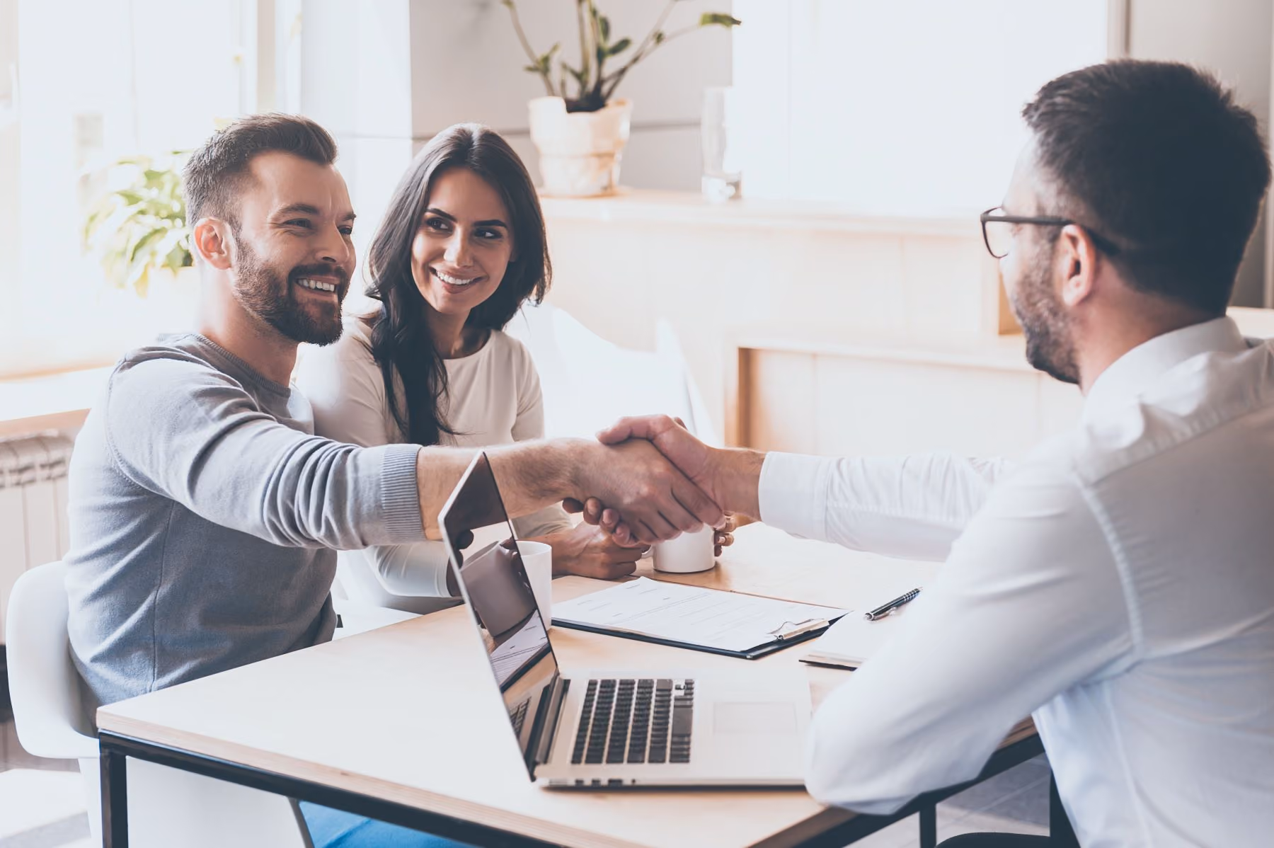 Man and woman smiling while shaking hands with another man across an office table with a laptop.