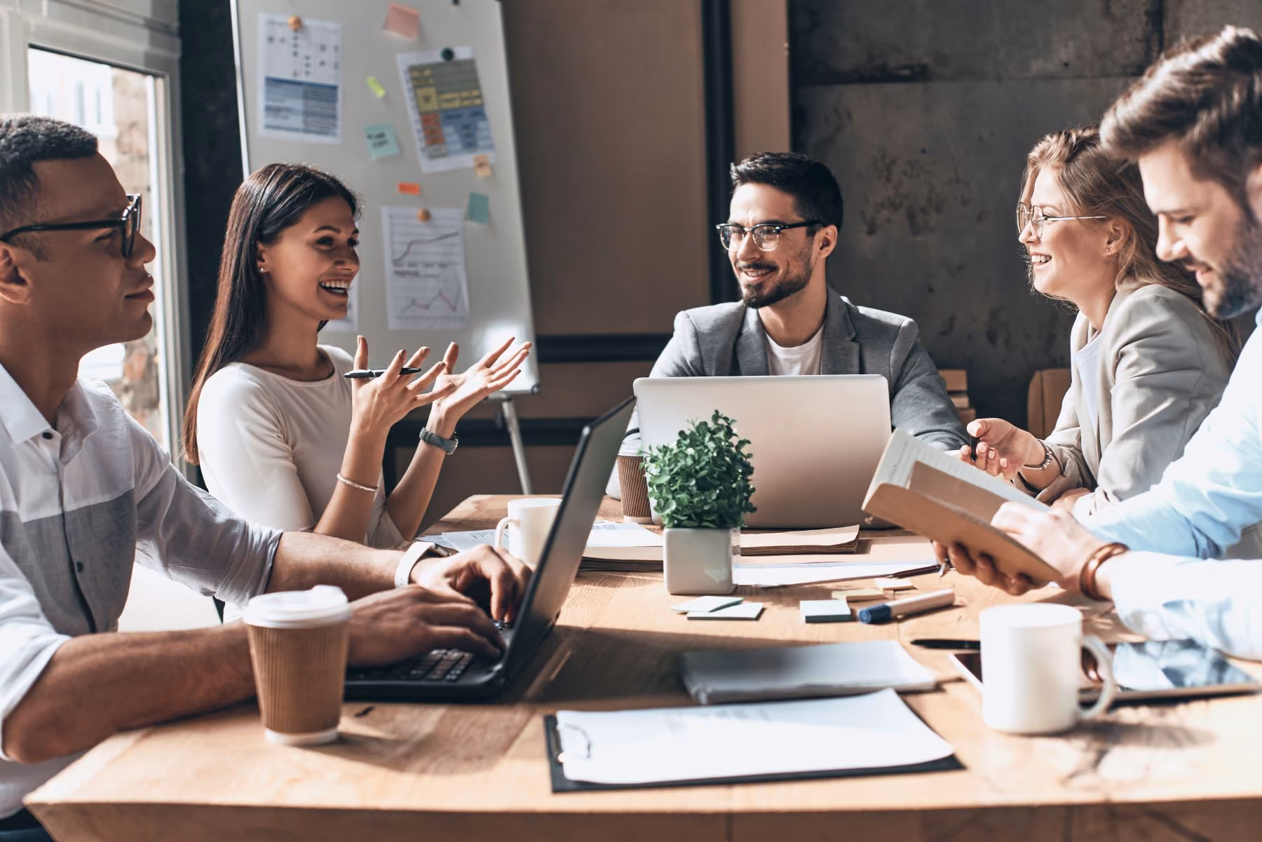 A team of lawyers collaborating around a table with laptops, papers, and coffee cups in a meeting room.