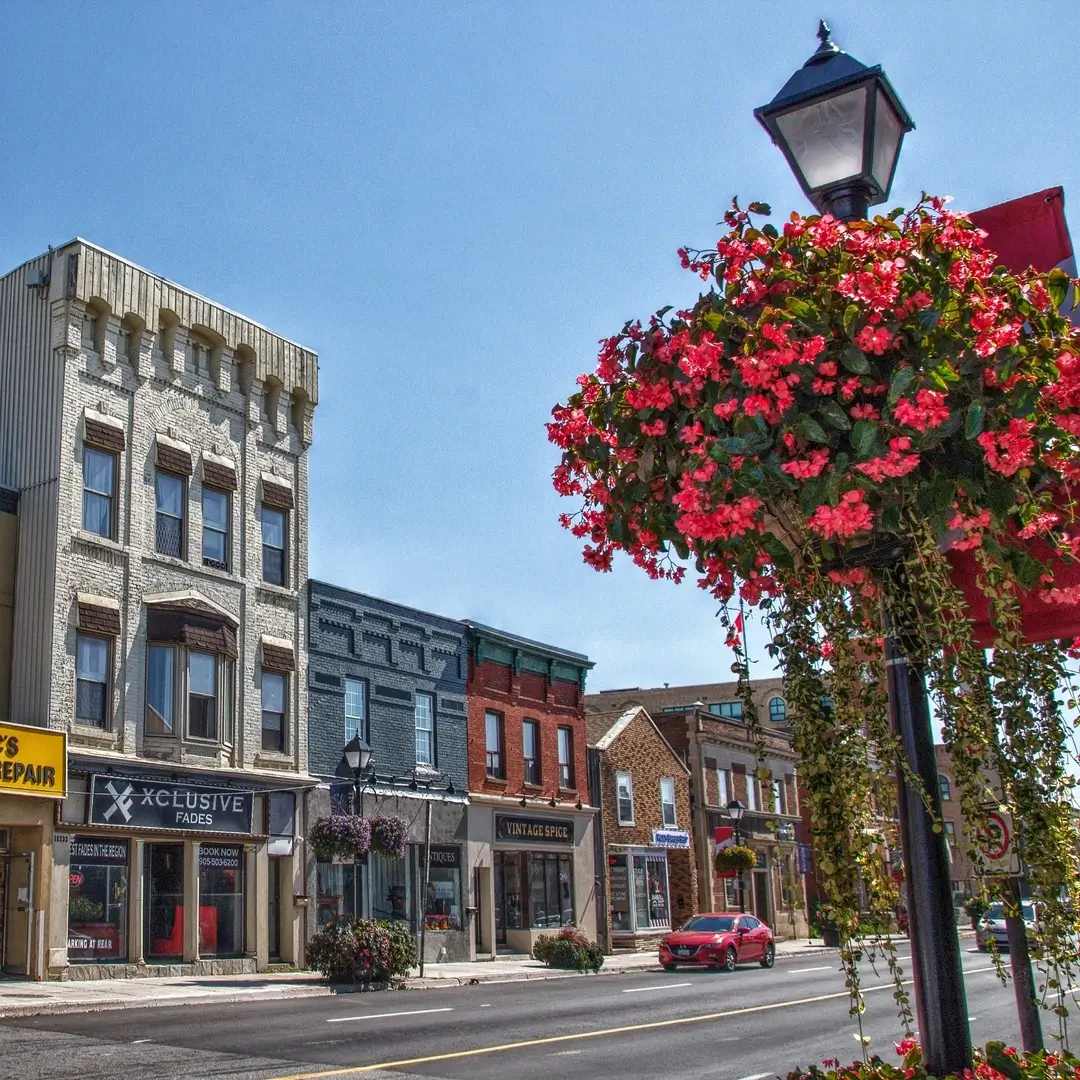 Street view of vintage storefronts with hanging flower baskets and a red car on a sunny day.