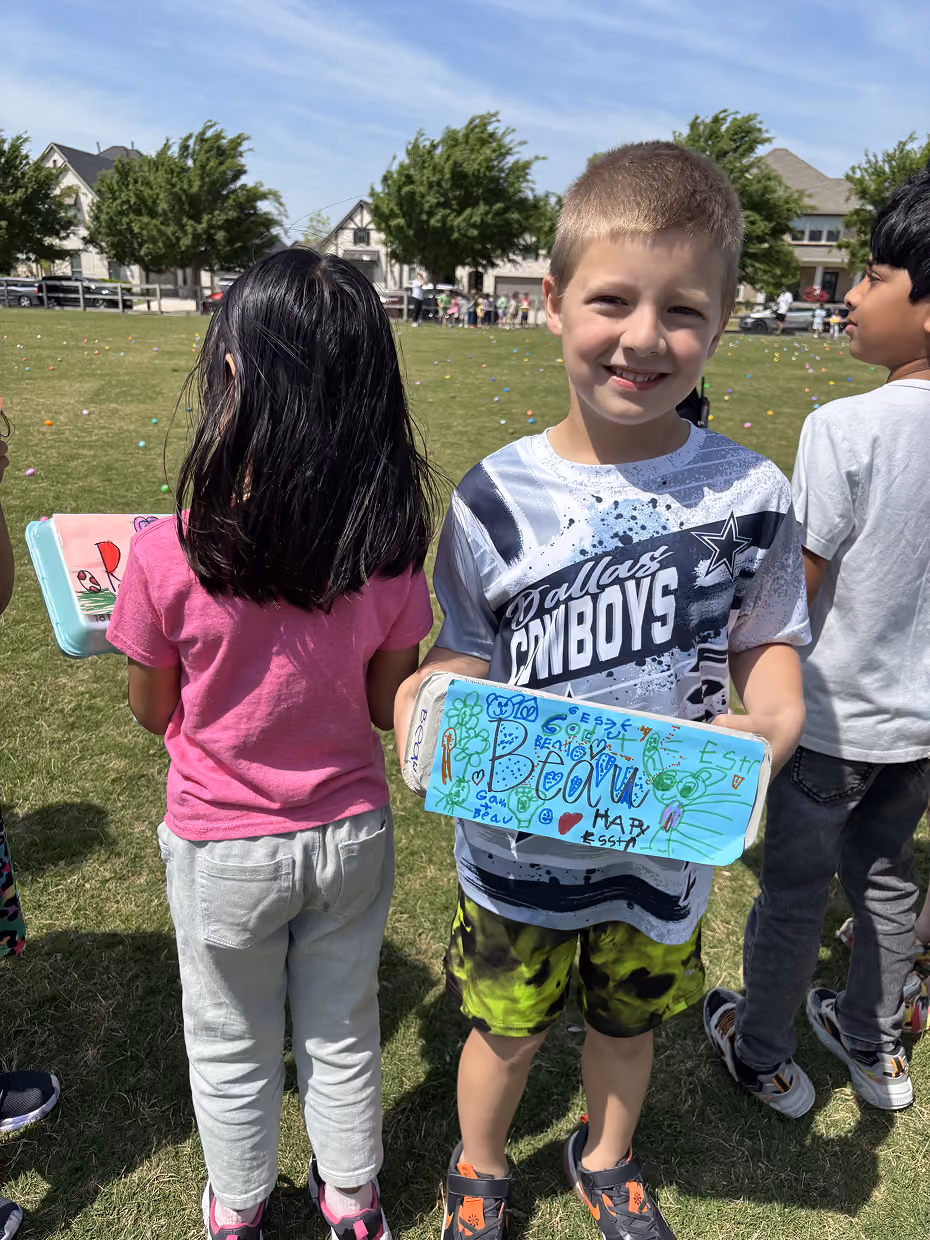 Smiling boy in Dallas Cowboys shirt holding decorated box with drawings and names on a grassy field.