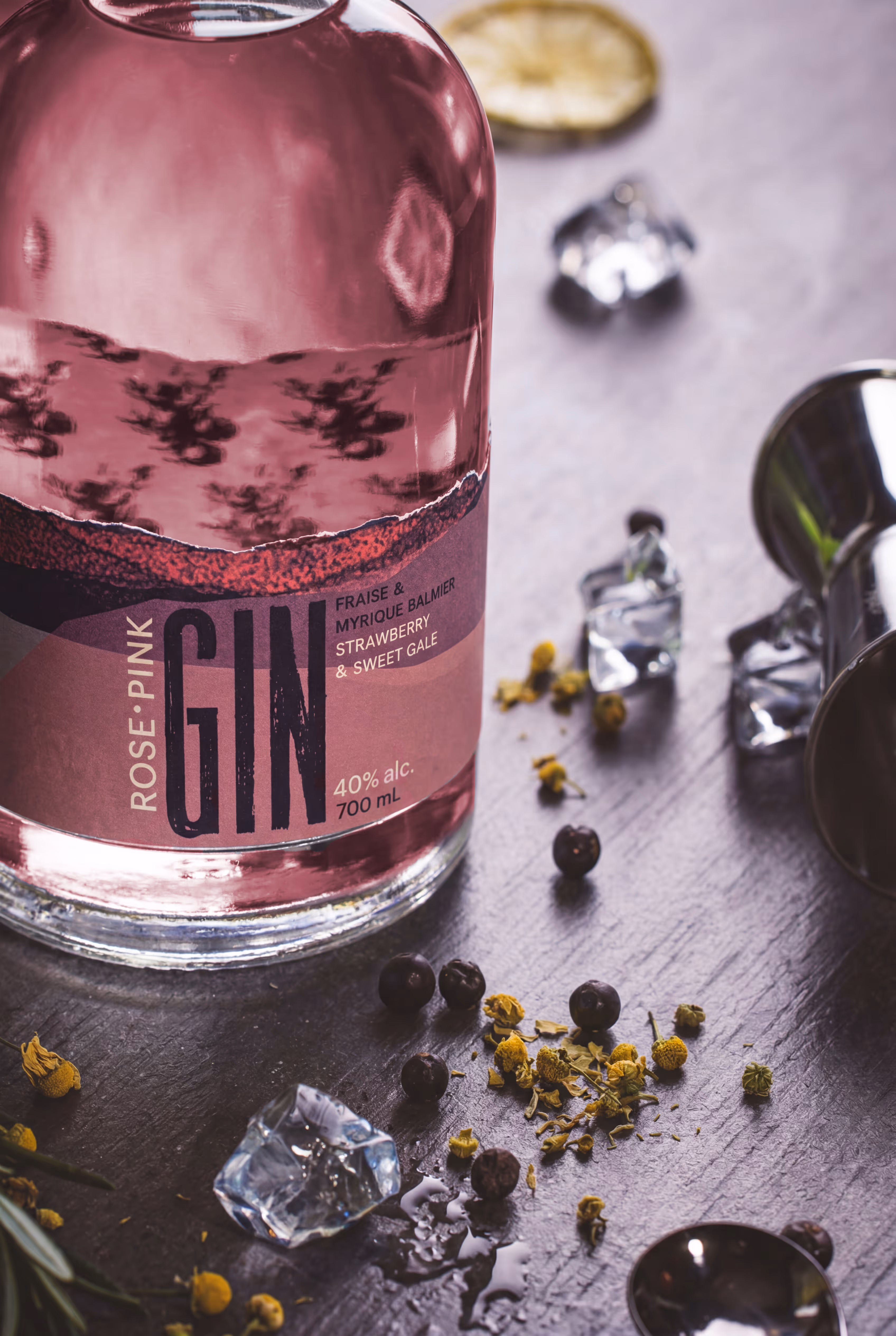 Close-up of a pink gin bottle with ice cubes, dried flowers, berries, and a jigger on a dark surface. (Adobe Firefly)