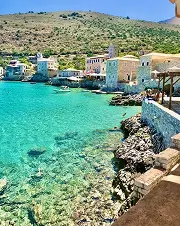 Clear turquoise sea along rocky shoreline with stone buildings and hills in the background.