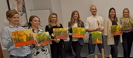 Seven people holding paintings of autumn trees, standing indoors in a row.
