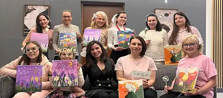 Group of 10 women holding up colorful floral paintings while posing indoors.