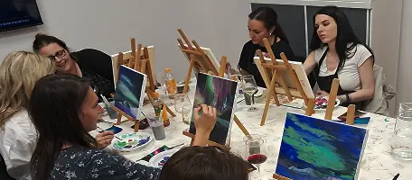 Group of women painting on canvases at a table in an art class or workshop.