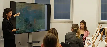 Woman presenting artwork on a screen to seated audience in a room with paintings on display.