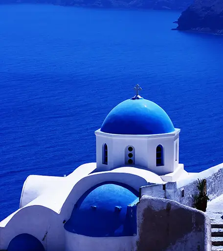 White building with two blue domes and a cross, overlooking deep blue sea and rocky coastline.