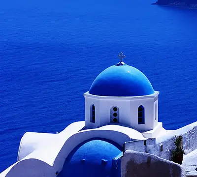 White building with blue domes and cross overlooking deep blue sea, typical of Santorini, Greece.