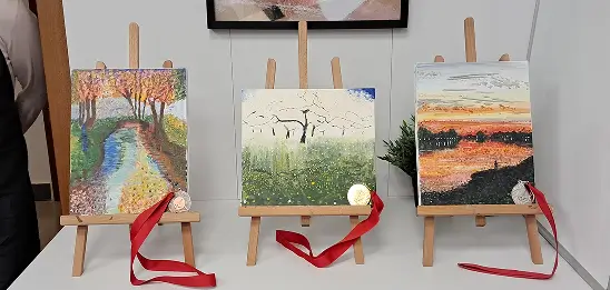 Three landscape paintings on wooden easels showing a river, flowering tree, and sunset scene with medals on red ribbons.
