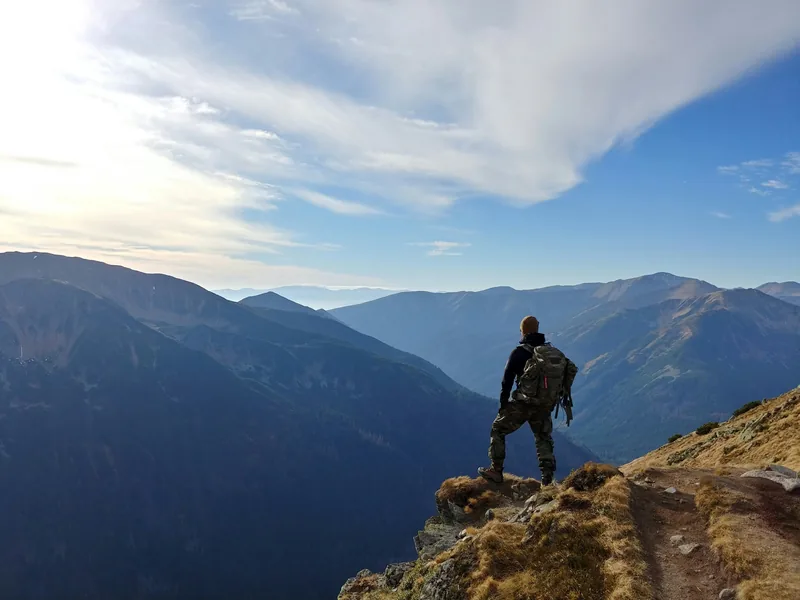 Hiker with backpack standing on rocky cliff overlooking mountain valley under a cloudy blue sky.