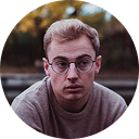 Young man with glasses and short light hair wearing a purple sweater outdoors.